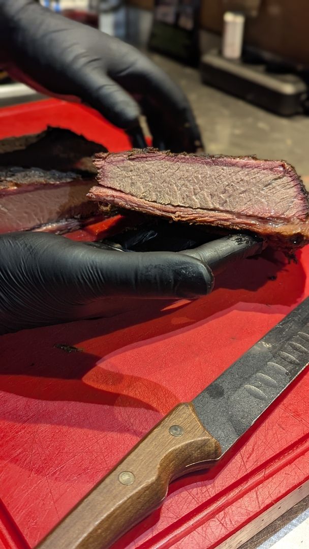 Eine Hand mit schwarzem Handschuh hält ein großes Stück geschnittenes Brisket mit einem rosa Rauchring auf einem roten Schneidebrett neben einem Messer.