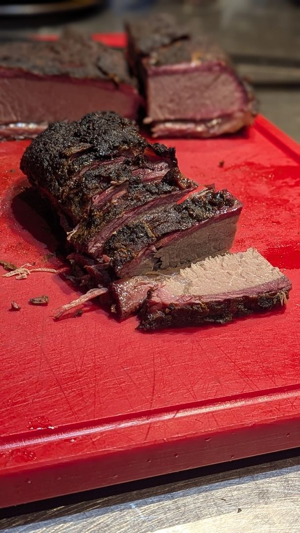 Mehrere Scheiben zartes Brisket mit einer dunklen Kruste und einem rosa Rauchring liegen auf einem roten Schneidebrett.